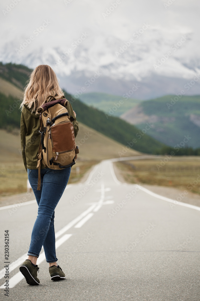 A woman with a backpack and a road stretching into the distance against the backdrop of mountains - obrazy, fototapety, plakaty 