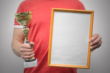 Wall Mural - Man is holding a blank diploma or certificate frame with copy space and golden award trophy in the hands isolated on gray background.