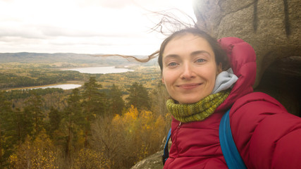 Girl in warm clothes with a scarf with a backpack on top of a rock mass makes a selfie on the background of autumn nature-a landscape with a lake. The brunette's hair is shaggy