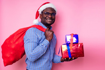 African American man wearing stylish plaid shirt great smile in santa hat with gift box on pink background studio.dark-skinned Santa Claus merry christmas with sack full of Christmas goodies