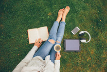 Top View Of Woman Sitting In Park (garden) On The Green Grass With Smartphone, Headphones, Tablet, Book And Coffee In The Hand