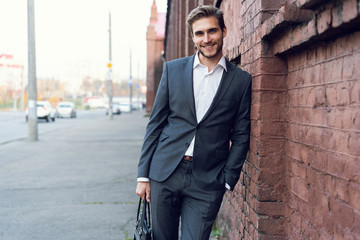 Smiling young male manager formal dressed leaning on a wall outdoors.