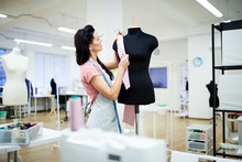 Serious Focused Brunette Young Lady In Apron Standing At Mannequin And Putting Fabric Piece While Working Alone In Dressmaking Studio