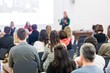 © kasto - Business and entrepreneurship symposium. Female speaker giving a talk at business meeting. Audience in conference hall. Rear view of unrecognized participant in audience. Copy space on whitescreen.