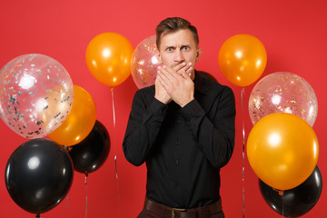 Shocked young man in black classic shirt covering mouth with palms on red background air balloons. St. Valentine's, International Women's Day, Happy New Year, birthday mockup holiday party concept.