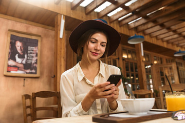 Poster - Portrait of young woman wearing hat using smartphone, while sitting in stylish cafe