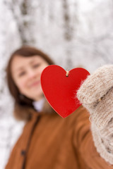 Happy girl with red wooden heart holding in hands in winter forest (unfocused background). Valentines day and Christmas holidays. Winter concept. Love and romantic background. Smiling girl with gloves