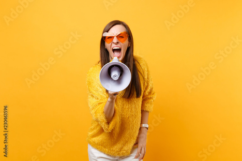 Photographie Portrait of crazy young woman in fur sweater and orange heart eyeglasses screaming on megaphone isolated on bright yellow background