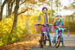 © MNStudio - Cute sisters riding bikes in a city park on sunny autumn day. Active family leisure with kids. Children wearing safety hemet while riding a bicycle.
