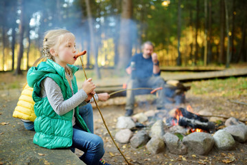 Wall Mural - cute little sisters and their father roasting hotdogs on sticks at bonfire. children having fun at c