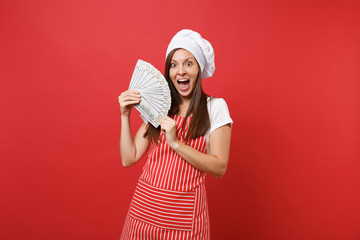 Housewife female chef cook baker in striped apron, white t-shirt, toque chefs hat isolated on red wall background. Happy woman holding lots of dollars banknotes cash money. Mock up copy space concept.