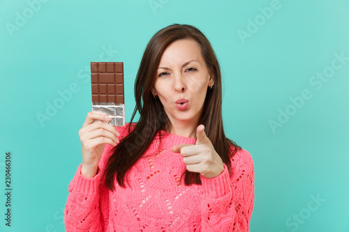 Fotografie, Tablou Stunning young woman in knitted pink sweater pointing index finger on camera hold in hand chocolate bar isolated on blue wall background, studio portrait