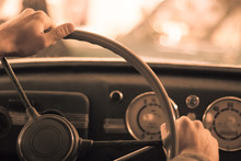 Driving A Vintage Car; Only The Driver’s Hand On The Steering Wheel Are Visible, The Dashboard Is Blurred; Stylized As An Old Sepia Photo With Dust And Noise