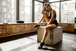 © Leika production - Charming slander girl dressed in black sports top and shorts is sitting on a wooden box in the sunlight in front the window in the gym