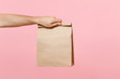Close up male holding in hand brown clear empty blank craft paper bag for takeaway isolated on pastel pink background. Packaging template mock up. Delivery service concept. Copy space advertising area