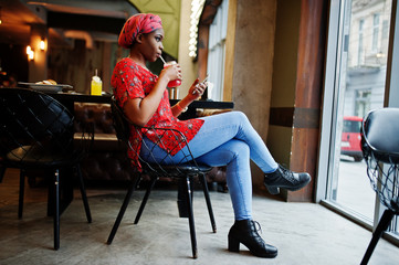 Wall Mural - Stylish african woman in red shirt and hat posed indoor cafe and drinking strawberry lemonade with phone at hand.