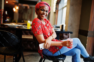 Wall Mural - Stylish african woman in red shirt and hat posed indoor cafe and drinking strawberry lemonade with phone at hand.