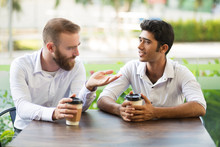 Two Male Friends Drinking Coffee And Chatting In Outdoor Cafe. People Sitting At Table With Blurred Plants In Background. Coffee Break Concept. Front View.