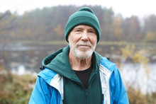 Outdoor Close Up Shot Of Handsome Active Elderly Male Pensioner With Gray Beard Having Morning Walk In Wild Nature, Posing Against Misty Lake And Colorful Autumn Forest Background, Looking At Camera