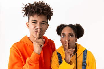 Image of african american brother and sister in colorful clothes holding fingers at mouth, isolated over white background