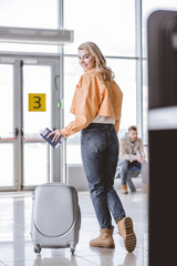 Wall Mural - attractive young woman with passport, boarding pass and suitcase smiling at camera in airport