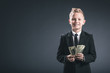 portrait of smiling preteen boy dressed as businessman holding dollar banknotes in hands on grey backdrop