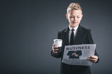 portrait of boy in businessman suit with coffee to go reading business newspaper on grey backdrop