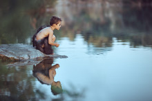 Caveman Boy Sitting On The Rock Near River Or Lake And Looking Away. Evolution Survival Concept. Creative Art Fantasy Photo