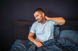 © estradaanton - Young man sits on bed and feels pain in neck area. He shrinks and looks to left. Guy holds hand on neck behind. He is partly covered with blanket.