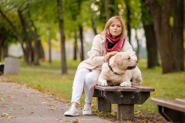 A young woman sits on a bench in the park, and her Labrador walks nearby.