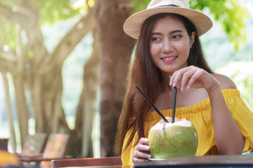 Happy smiling face traveler young women wearing yellow dress with hat drinking fresh coconut water in restaurant in the garden.