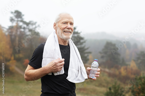 Exhausted elderly male with gray hair and beard drinking water after outdoor exercise and wiping sweat with towel around his neck. Tired but happy old senior man resting after running workout