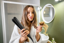Portrait Of Unhappy Woman Holding Splitting Hair Ends And Hair Straightener In The Bathroom