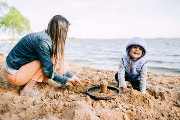 Mother and daughter playing together. Young female with her child building castle in sand on beach near sea. Parent with funny baby lifestyle portrait. Motherhood and childhood. Happy family outdoor.