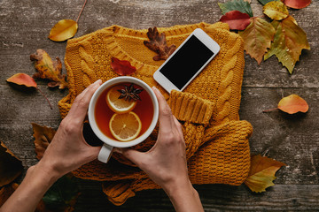 Wall Mural - partial view of woman holding cup of tea on wooden tabletop with knitted sweater, smartphone and fallen leaves