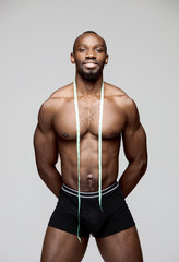 Fit young man with measuring tape isolated on gray background. The naked torso of African American man posing at studio. The muscular body, fitness, sports, healthy lifestyle and bodybuilder concept.