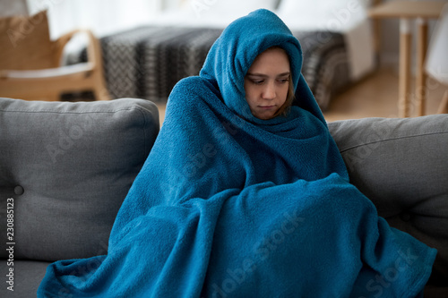 Fotomural Young unsatisfied sick woman covered with warm blue blanket plaid from head to toes sitting alone on couch in living room at cold home