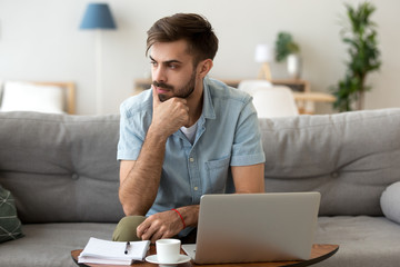 Contemplating millennial serious man looking away sitting on couch. Thoughtful pensive handsome serious male student thinking about new idea or project analysing planning making decision concept