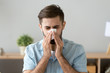 Young sick man sitting at office room sneeze holding tissue handkerchief blowing wiping his running nose. Headshot Male feels unwell caught influenza cold seasonal infection or having allergy concept