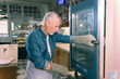 © zinkevych - Near oven. Grey-haired owner of bakery wearing denim shirt standing near oven looking at baguettes