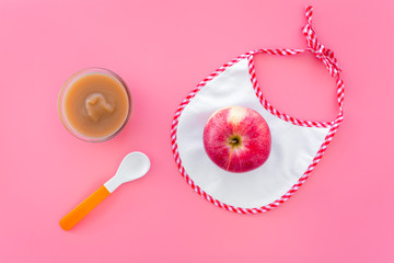 Baby food concept. Apple puree in bowl near bib, apple, spoon on pink background top view