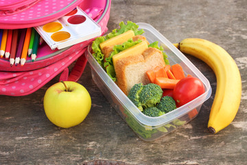 Sandwiches, fruits and vegetables in food box, backpack on old wooden background.