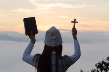 Wall Mural -  Young woman hands holding holy Bible and lift of christian cross with light sunset background.