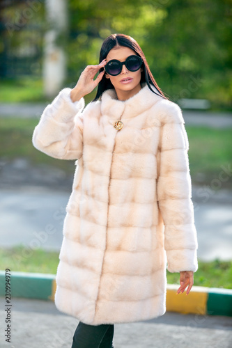 Fotografia, Obraz Beautiful brunette in sunglasses and a light fur coat walking down the street