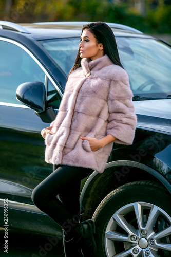 Fotografie, Obraz A beautiful brunette in a light fur coat and black trousers is standing near the