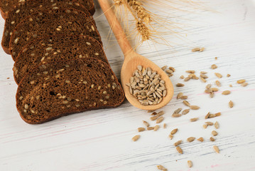 Rural still life on a white wooden background - sliced rye brown bread, sunflower ,seeds on a light  spoon and ears of wheat