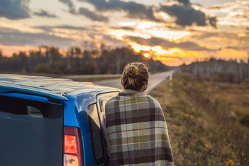 A woman in a plaid stands by the car on the side of the road in the background of the dawn. Road trip concept