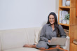 © DragonImages - Beautiful smiling Asian woman sitting barefoot on couch with laptop smiling at camera in modern office