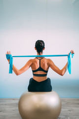 Young sporty attractive woman practicing pilates with resistance bands back sitting on fitness balls on white urban studio. Yoga and Pilates lifestyle healthy concept.