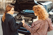 © LIGHTFIELD STUDIOS - high angle view of curly ginger woman giving wrench to female friend in coat near car with opened hood at city street